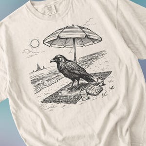 May include: Cream-colored t-shirt featuring a black and white illustration of a raven on a beach. The raven is under a beach umbrella, with a soda can and a bag of potato chips nearby. The shirt has a Comfort Colors tag.