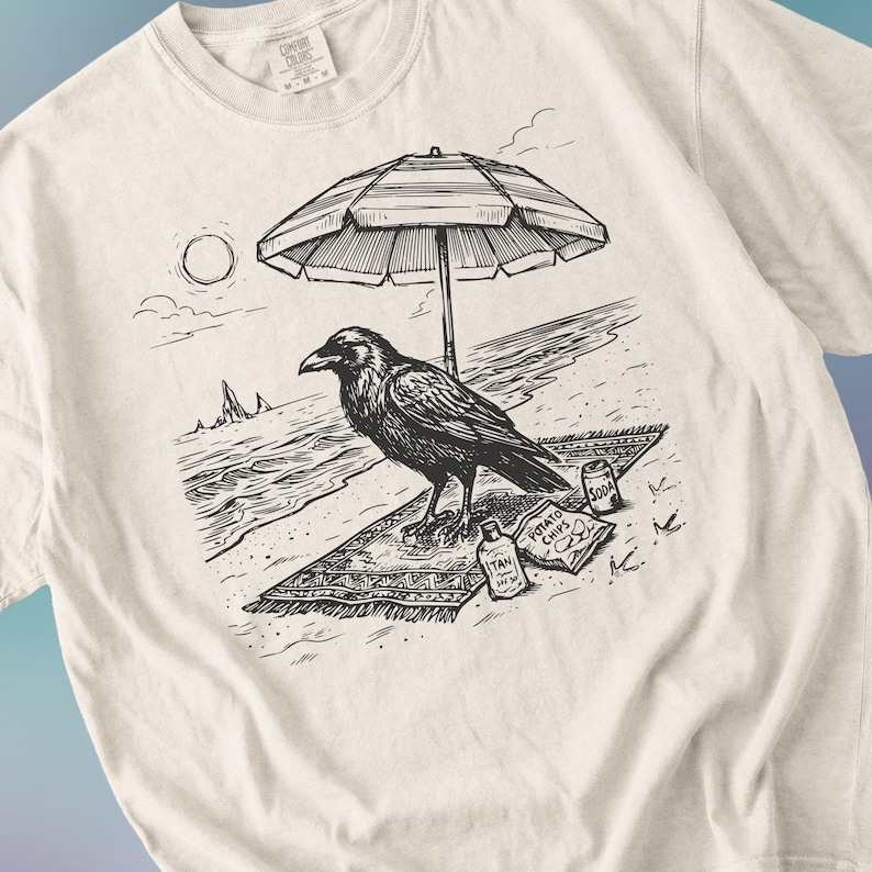 May include: Cream-colored t-shirt featuring a black and white illustration of a raven on a beach. The raven is under a beach umbrella, with a soda can and a bag of potato chips nearby. The shirt has a Comfort Colors tag.