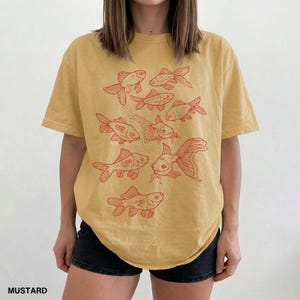 May include: A mustard yellow short-sleeve t-shirt featuring a graphic print of multiple goldfish in a reddish-orange ink. The design consists of several fish illustrations arranged in a cluster across the front of the garment, which is paired with black shorts.