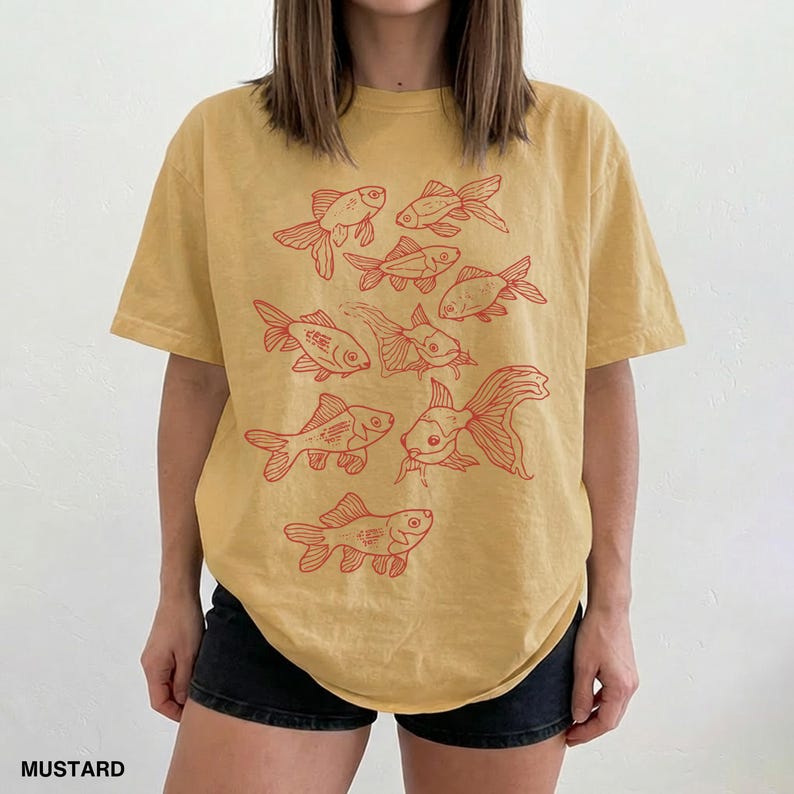 May include: A mustard yellow short-sleeve t-shirt featuring a graphic print of multiple goldfish in a reddish-orange ink. The design consists of several fish illustrations arranged in a cluster across the front of the garment, which is paired with black shorts.