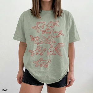 May include: A sage green short-sleeve t-shirt featuring a graphic print of multiple goldfish in red ink. The design displays various fish swimming in different directions across the front of the garment, which is paired with black shorts.