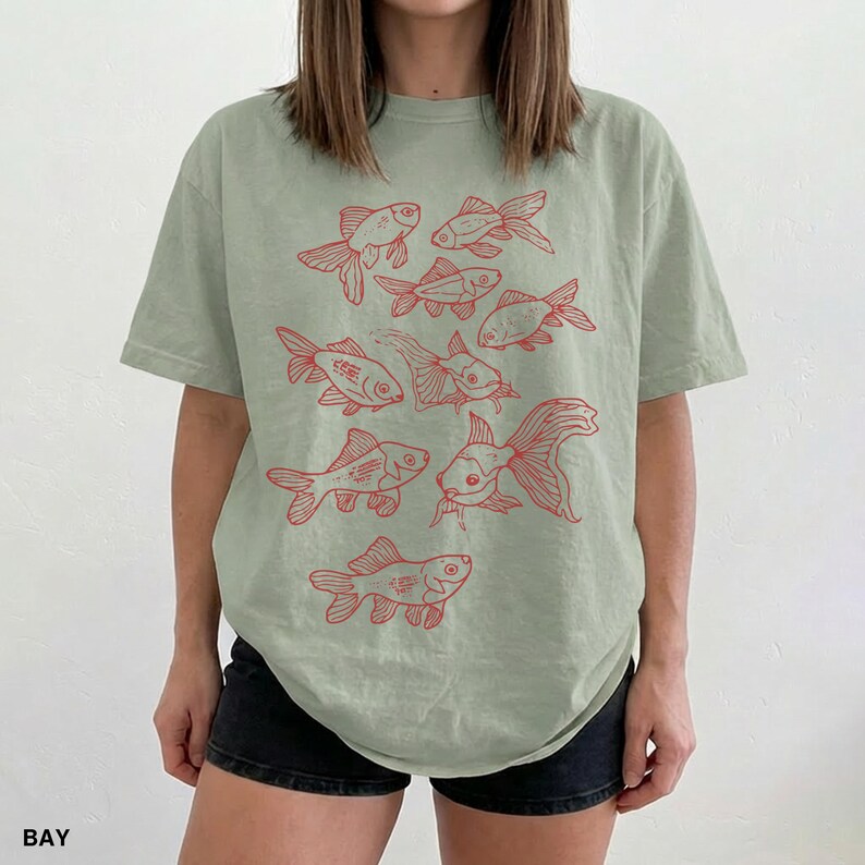 May include: A sage green short-sleeve t-shirt featuring a graphic print of multiple goldfish in red ink. The design displays various fish swimming in different directions across the front of the garment, which is paired with black shorts.