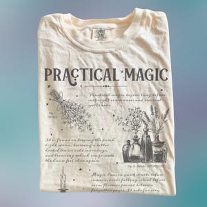Puede incluir: Camiseta color crema con las palabras "PRACTICAL MAGIC" en gris oscuro. La camiseta presenta ilustraciones de hierbas, botellas y texto que describe la magia práctica. La camiseta está doblada.