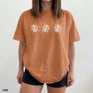 May include: A burnt orange short-sleeve t-shirt featuring a horizontal row of three white line-art illustrations of ladybugs centered on the chest. The shirt has a crew neckline and a relaxed fit, providing a casual look for everyday wear.