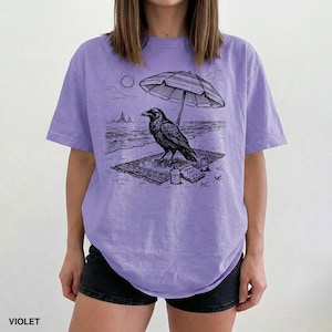 May include: Violet-colored t-shirt featuring a black and white illustration of a raven on a beach under an umbrella. The raven is on a rug with a bottle of sunscreen and a bag of chips. The word "VIOLET" is printed at the bottom.