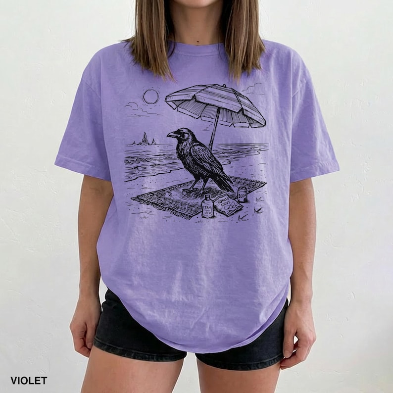 May include: Violet-colored t-shirt featuring a black and white illustration of a raven on a beach under an umbrella. The raven is on a rug with a bottle of sunscreen and a bag of chips. The word "VIOLET" is printed at the bottom.