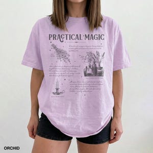 May include: A light purple t-shirt with the words "PRACTICAL MAGIC" in a stylized font. The shirt features illustrations of herbs, candles, and bottles with text describing magical practices. The word "ORCHID" is printed at the bottom.