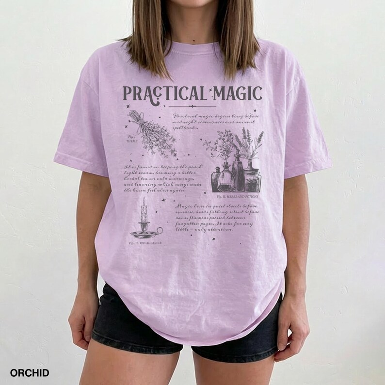 May include: A light purple t-shirt with the words "PRACTICAL MAGIC" in a stylized font. The shirt features illustrations of herbs, candles, and bottles with text describing magical practices. The word "ORCHID" is printed at the bottom.