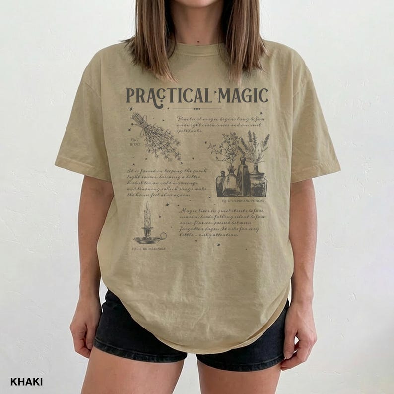 May include: Khaki-colored t-shirt with the words "PRACTICAL MAGIC" printed on the front. The design includes illustrations of herbs, bottles, and a candle. The shirt has a relaxed fit and short sleeves.