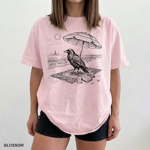 May include: Light pink t-shirt with a black and white graphic of a raven on a beach, under an umbrella. The design includes the sun, waves, and the text "BLOSSOM" at the bottom.