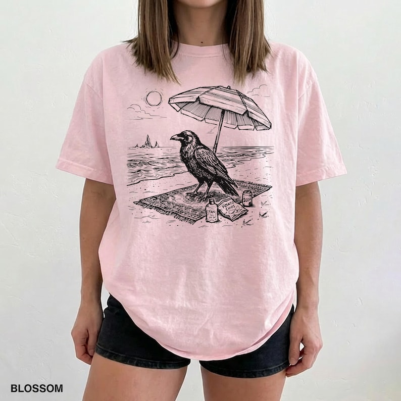 May include: Light pink t-shirt with a black and white graphic of a raven on a beach, under an umbrella. The design includes the sun, waves, and the text "BLOSSOM" at the bottom.