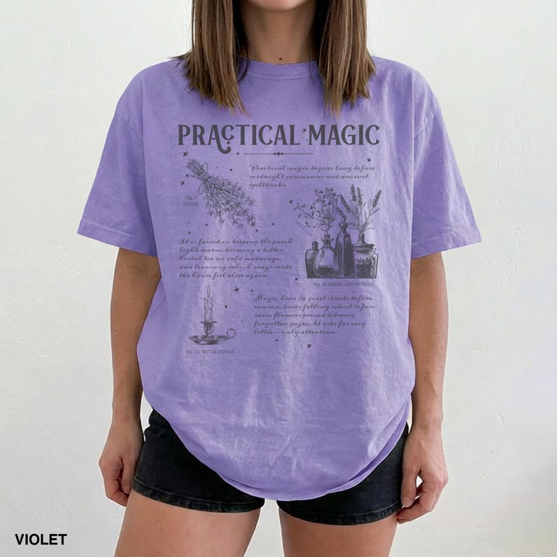 May include: Violet-colored t-shirt with the text "PRACTICAL MAGIC" and botanical illustrations. The shirt features a crew neck and short sleeves. The design includes images of herbs, candles, and potion bottles.