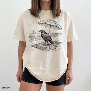 May include: Ivory-colored t-shirt featuring a black and white graphic of a raven on a beach, under an umbrella. The design includes the sun, waves, and small bottles. The shirt is a relaxed fit.