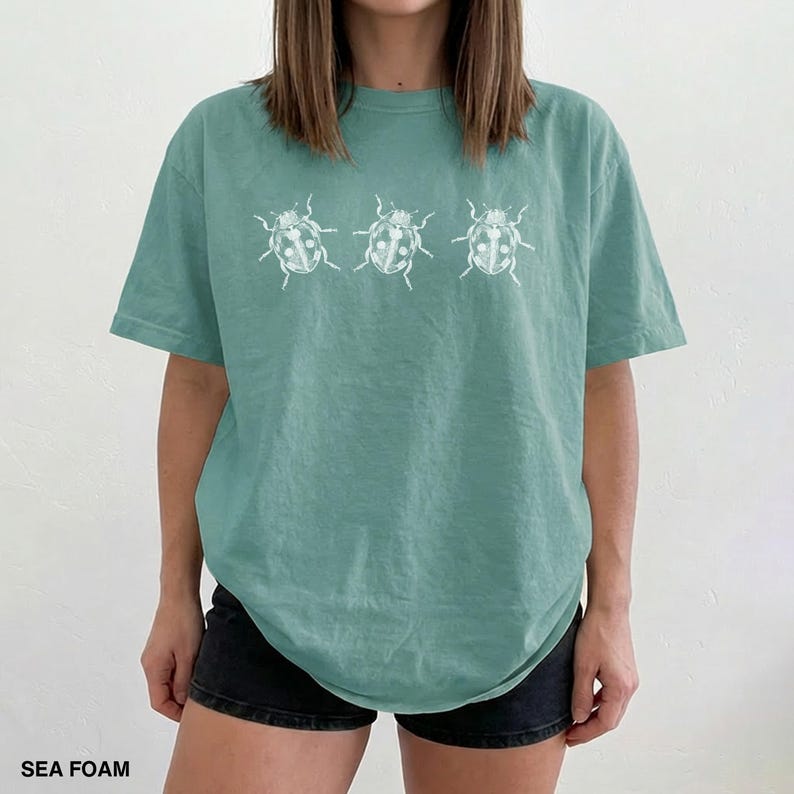 May include: A sea foam green short-sleeve t-shirt featuring a white graphic print of three ladybugs arranged in a horizontal row across the chest. The shirt has a crew neckline and a relaxed fit, providing a casual look with a nature-inspired design.