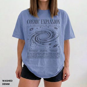 May include: A light blue t-shirt with the text "COSMIC EXPANSION" and a graphic of a galaxy. The shirt also has additional text describing the expansion of the universe. The shirt is made of washed denim.