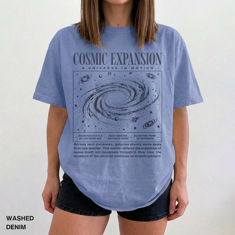 May include: A light blue t-shirt with the text "COSMIC EXPANSION" and a graphic of a galaxy. The shirt also has additional text describing the expansion of the universe. The shirt is made of washed denim.