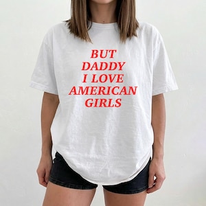 Könnte beinhalten: Weißes T-Shirt mit rotem Text "BUT DADDY I LOVE AMERICAN GIRLS". Das kurzärmlige Shirt hat eine lockere Passform. Der Text ist mittig auf der Vorderseite platziert.
