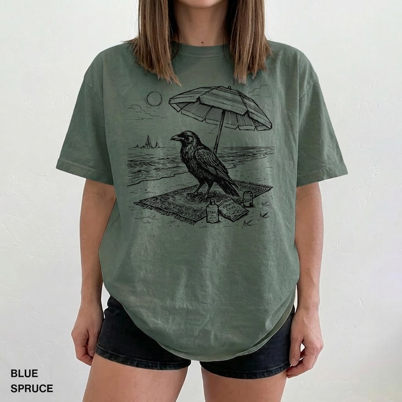 May include: Blue spruce t-shirt featuring a black and white graphic of a raven on a beach under an umbrella. The raven is perched on a rug with a bottle and book. The text "BLUE SPRUCE" is at the bottom.