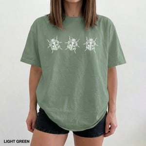 May include: A light green short-sleeved t-shirt featuring a white graphic print of three ladybugs arranged in a horizontal row across the chest. The shirt has a crew neckline and a relaxed fit, constructed from a solid-colored cotton fabric.