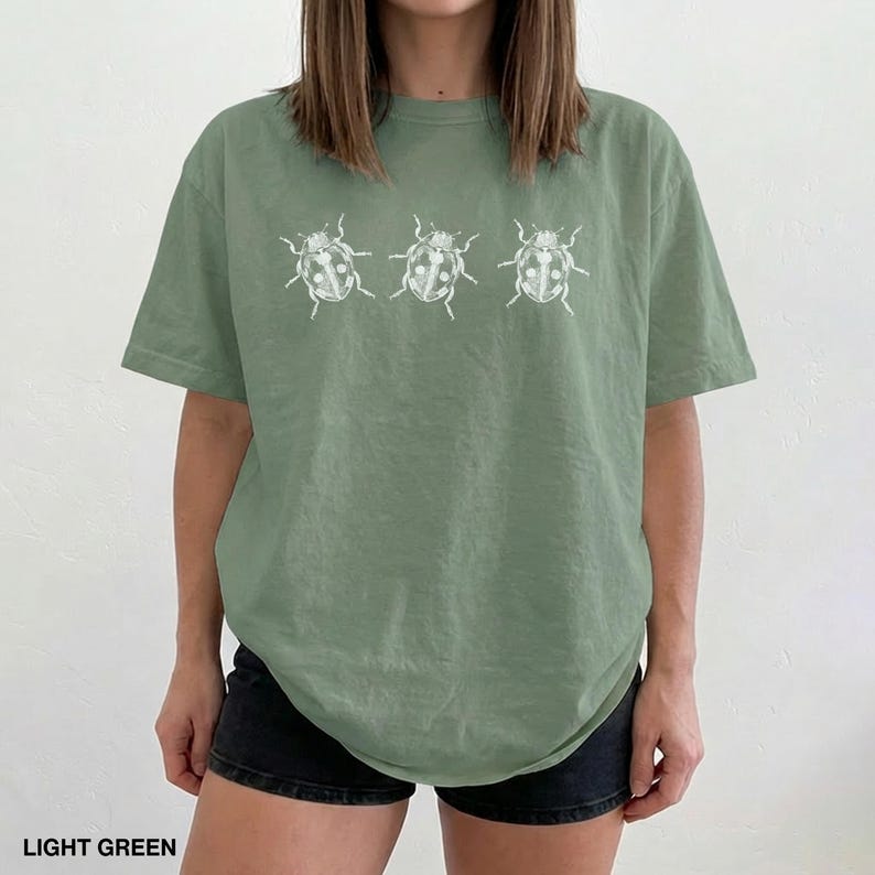 May include: A light green short-sleeved t-shirt featuring a white graphic print of three ladybugs arranged in a horizontal row across the chest. The shirt has a crew neckline and a relaxed fit, constructed from a solid-colored cotton fabric.