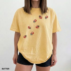 Może przedstawiać: Ż&oacute;łty t-shirt z okrągłym dekoltem z wzorem czerwonych i czarnych biedronek. Koszulka ma jasny ż&oacute;łty odcień i lekko wyblakły wygląd. Słowo "BUTTER" jest wydrukowane w lewym dolnym rogu obrazu.