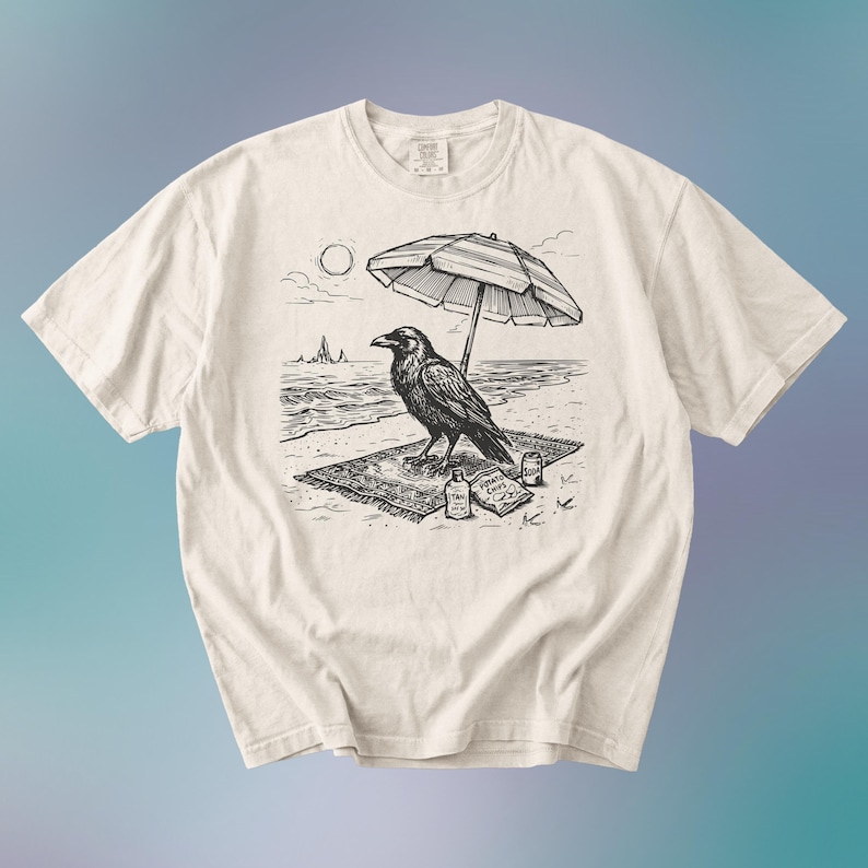 May include: Cream-colored t-shirt featuring a black and white illustration of a raven relaxing on a beach. The raven is under a striped umbrella, with the sun, ocean, and snacks visible. A unique graphic tee.