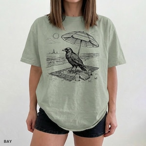 May include: Sage green t-shirt featuring a black and white illustration of a raven relaxing on a beach. The raven is under a striped umbrella, with the sun and ocean in the background. The word "BAY" is printed at the bottom left.