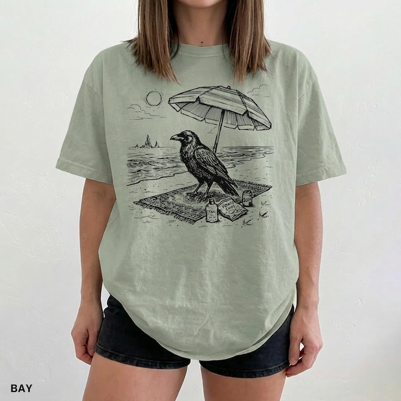 May include: Sage green t-shirt featuring a black and white illustration of a raven relaxing on a beach. The raven is under a striped umbrella, with the sun and ocean in the background. The word "BAY" is printed at the bottom left.