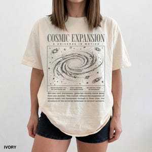 May include: Ivory-colored t-shirt with a graphic of a galaxy and the text "COSMIC EXPANSION - A UNIVERSE IN MOTION." The design includes text about the universe's expansion. The shirt is a casual, unisex style.