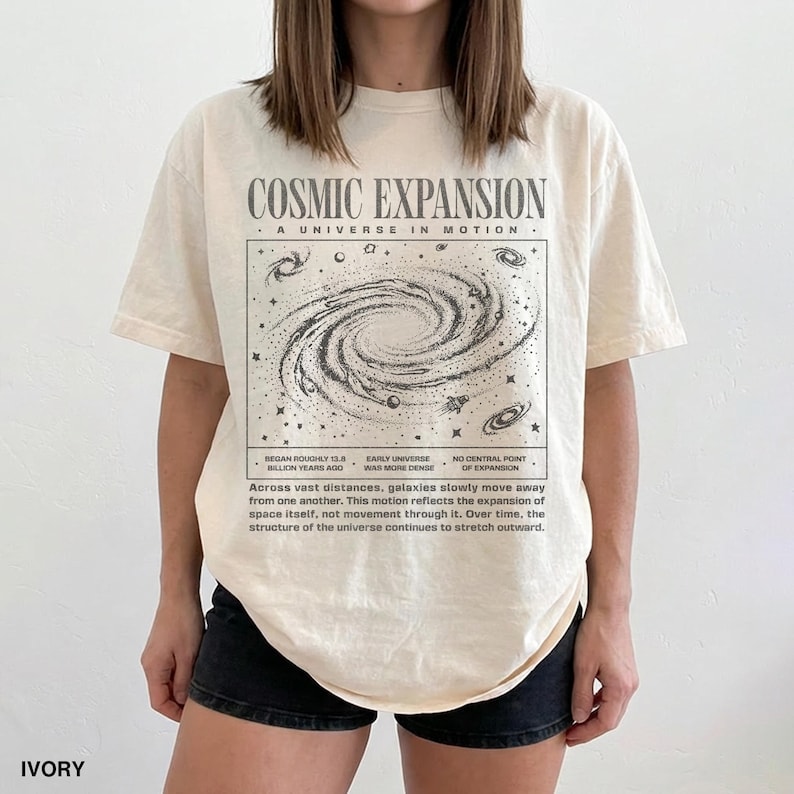 May include: Ivory-colored t-shirt with a graphic of a galaxy and the text "COSMIC EXPANSION - A UNIVERSE IN MOTION." The design includes text about the universe's expansion. The shirt is a casual, unisex style.