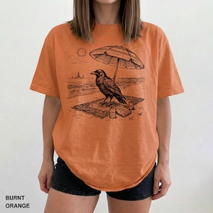 May include: Burnt orange t-shirt featuring a black graphic of a raven relaxing on a beach under an umbrella. The raven is on a rug with snacks and drinks. The shirt has the words "BURNT ORANGE" at the bottom.