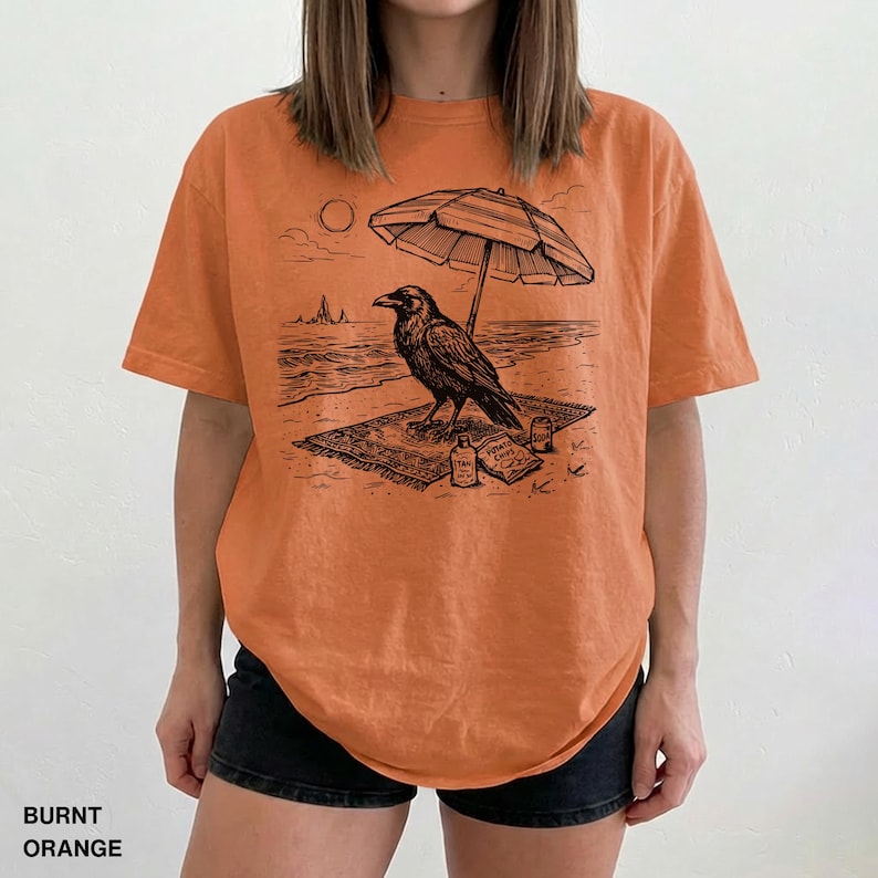 May include: Burnt orange t-shirt featuring a black graphic of a raven relaxing on a beach under an umbrella. The raven is on a rug with snacks and drinks. The shirt has the words "BURNT ORANGE" at the bottom.