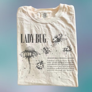 Könnte beinhalten: Cremefarbenes T-Shirt mit dem Text "LADY BUG" und Illustrationen von Marienkäfern und einer Blume. Das Shirt enthält auch den Text "MINIATURE CONSTELLATIONS" und zusätzlichen beschreibenden Text über Marienkäfer.