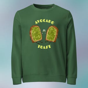 Pode incluir: Camisola verde escuro com um gráfico de duas fatias de torrada de abacate com braços e pernas, segurando taças de champanhe. As palavras "AVOCADO TOAST" estão em amarelo acima e abaixo da torrada.