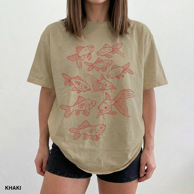 May include: A khaki t-shirt featuring a graphic print of multiple goldfish in red ink. The design consists of several fish illustrations arranged across the front of the shirt, showcasing various swimming positions and fin details in a line art style.
