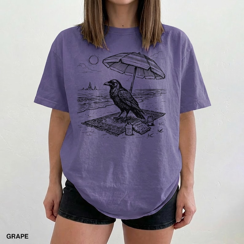 May include: Grape-colored t-shirt featuring a black and white illustration of a raven relaxing on a beach under an umbrella. The design includes a beach towel, a bottle, and a book. The word "GRAPE" is printed at the bottom.