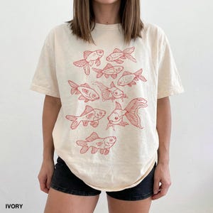 May include: An ivory short-sleeve t-shirt featuring a red line-art graphic print of multiple goldfish swimming in various directions across the front. The shirt has a crew neckline and a relaxed fit, showcasing a repeating pattern of fish.