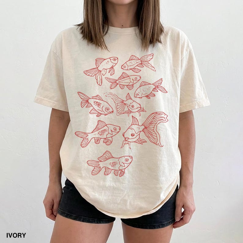 May include: An ivory short-sleeve t-shirt featuring a red line-art graphic print of multiple goldfish swimming in various directions across the front. The shirt has a crew neckline and a relaxed fit, showcasing a repeating pattern of fish.