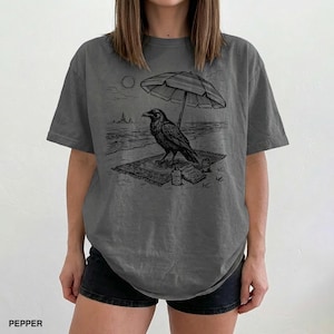 May include: Dark grey t-shirt featuring a black and white illustration of a raven on a beach under an umbrella. The raven is sitting on a rug with a bottle and book. The word "PEPPER" is printed at the bottom.