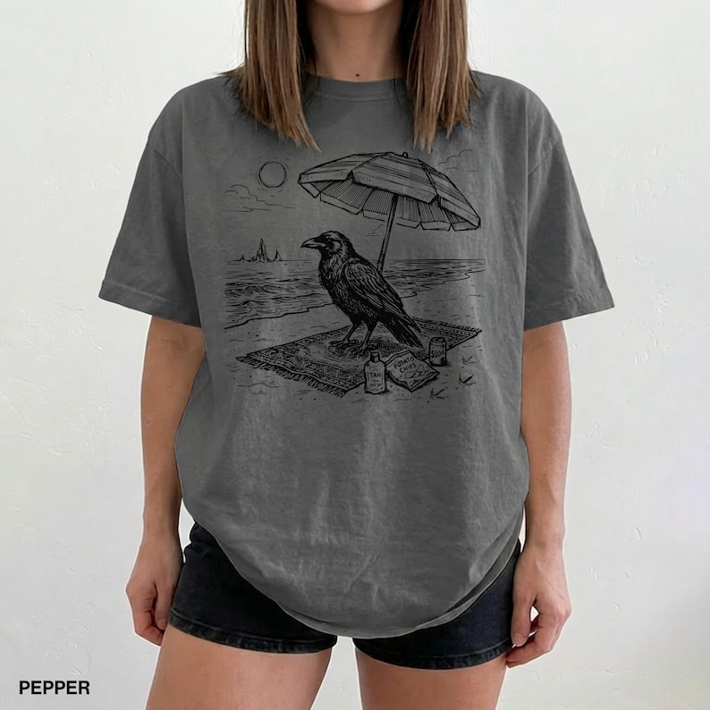May include: Dark grey t-shirt featuring a black and white illustration of a raven on a beach under an umbrella. The raven is sitting on a rug with a bottle and book. The word "PEPPER" is printed at the bottom.