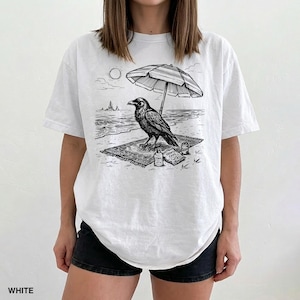 May include: White t-shirt featuring a black and white illustration of a raven relaxing on a beach. The raven is under a striped umbrella, with the sun, waves, and snacks visible. The word "WHITE" is printed at the bottom.