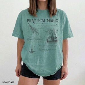May include: Sea foam green t-shirt with the words "PRACTICAL MAGIC" and botanical illustrations. The shirt features a design with herbs, bottles, and a candle, with text describing practical magic.