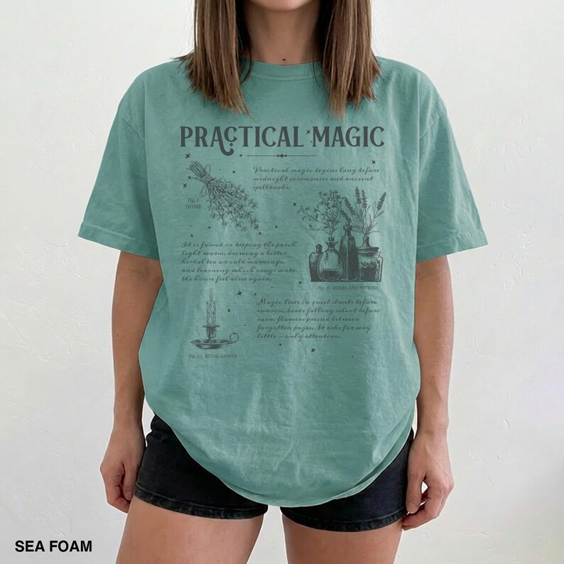 May include: Sea foam green t-shirt with the words "PRACTICAL MAGIC" and botanical illustrations. The shirt features a design with herbs, bottles, and a candle, with text describing practical magic.