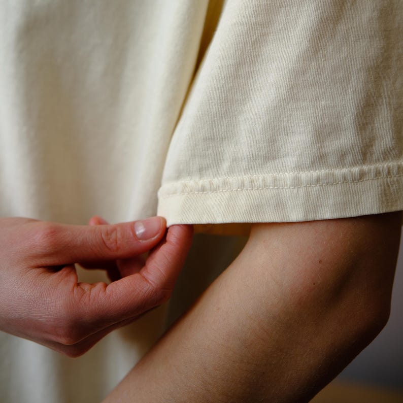May include: A short-sleeved t-shirt in a solid pale cream color. The garment features a standard crew neckline and a straight hemmed sleeve with visible double-needle stitching along the cuff edge, showcasing the texture of the fabric.