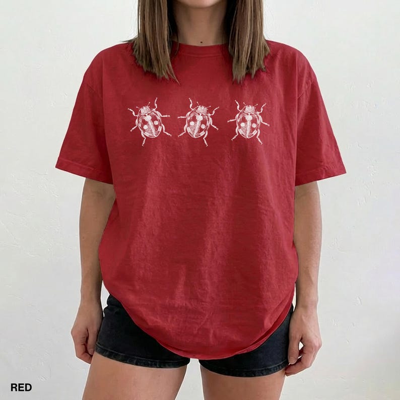 May include: A red short-sleeved t-shirt featuring a white graphic print of three ladybugs arranged in a horizontal row across the chest. The shirt has a crew neckline and a relaxed fit, with the ladybug illustrations showing detailed line work.
