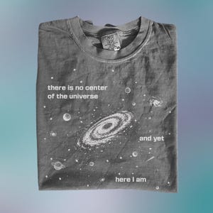 Op de afbeelding: Grijs T-shirt met een ronde hals en de tekst "there is no center of the universe" en "and yet here I am" met een galaxy-ontwerp. Het shirt is opgevouwen en heeft een label.
