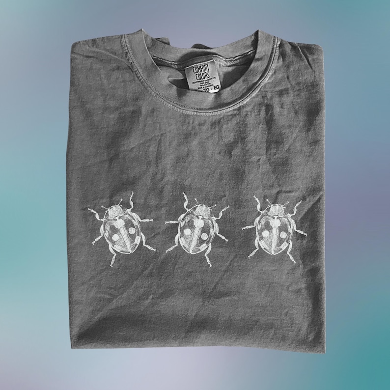 May include: A charcoal gray short-sleeve t-shirt featuring a white graphic print of three ladybugs arranged in a horizontal row across the center. The shirt has a crew neckline and a Comfort Colors brand label printed on the inside of the collar.