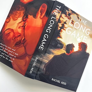 Puede incluir: Un libro titulado "The Long Game" de Rachel Reid. La portada presenta dos figuras en una paleta de colores cálidos, con el título y el nombre del autor en blanco. El lomo del libro también muestra el título.