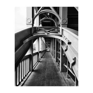 May include: Monochrome photograph of a bridge walkway. The image depicts the interior of a metal bridge with arched supports and a long, textured walkway. The perspective draws the eye along the length of the bridge.