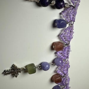 Puede incluir: Un rosario con una serie de cuentas de piedras preciosas de colores en tonos morados, verdes y marrones, conectadas por eslabones plateados. Las cuentas están ensartadas a lo largo de un delicado encaje de ganchillo lavanda.